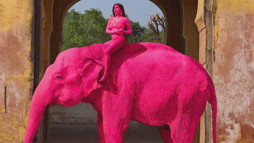 A bright pink painted elephant in an Indian city, surrounded by people.